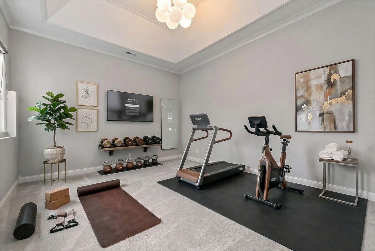 Just Listed! This Brentwood Estate Was Designed for Gathering, Entertaining & Everyday Living - 17 A home gym at Brentwood Estate with a treadmill, stationary bike, yoga mat, free weights, exercise accessories, a wall-mounted TV for entertaining workouts, framed art, and a potted plant on light carpeted flooring.
