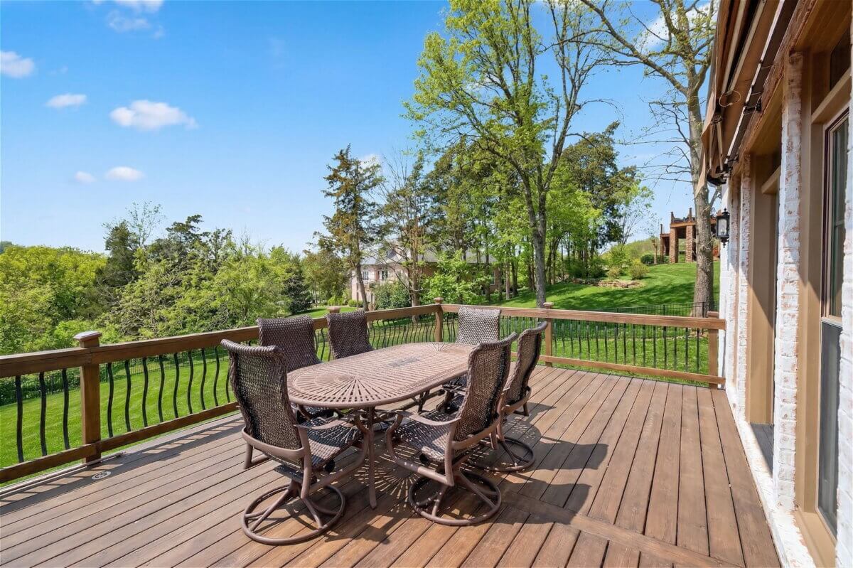 Just Listed! This Brentwood Estate Was Designed for Gathering, Entertaining & Everyday Living - 8 Outdoor wooden deck at Brentwood Estate with a metal dining table and six chairs, perfect for entertaining or everyday living, overlooking a green lawn and trees under a blue sky.