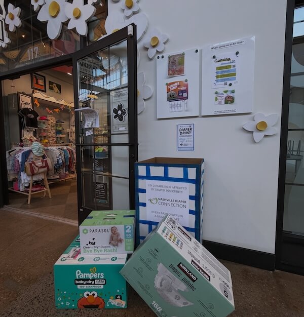 Your Guide to Nashville's Coolest Children's Stores - 2 Boxes of diapers and wipes are stacked near a donation bin outside a Nashville children’s store, with informational posters and decorative flowers on the wall.