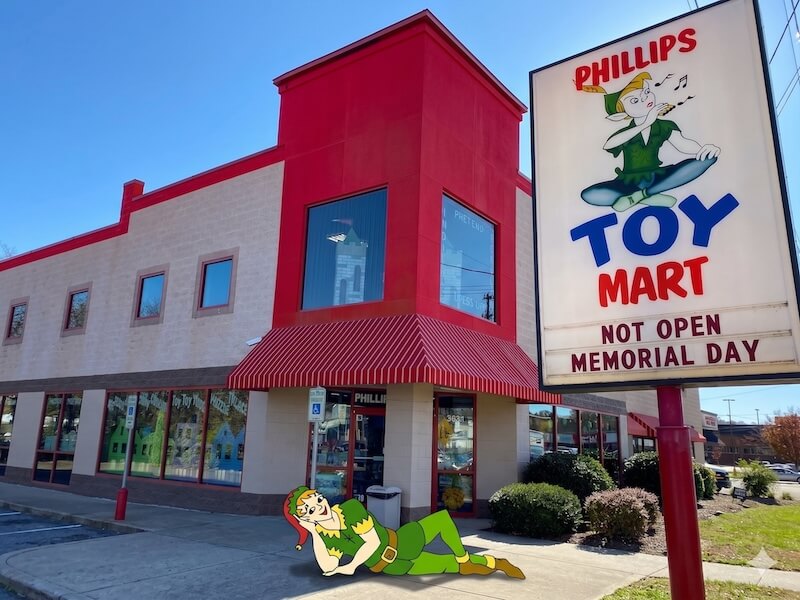 Your Guide to Nashville's Coolest Children's Stores - 4 A red and beige building in Nashville with a sign reading "Phillips Toy Mart, Not Open Memorial Day," features a cartoon elf character on the sign and sidewalk, capturing the charm of classic children's stores.