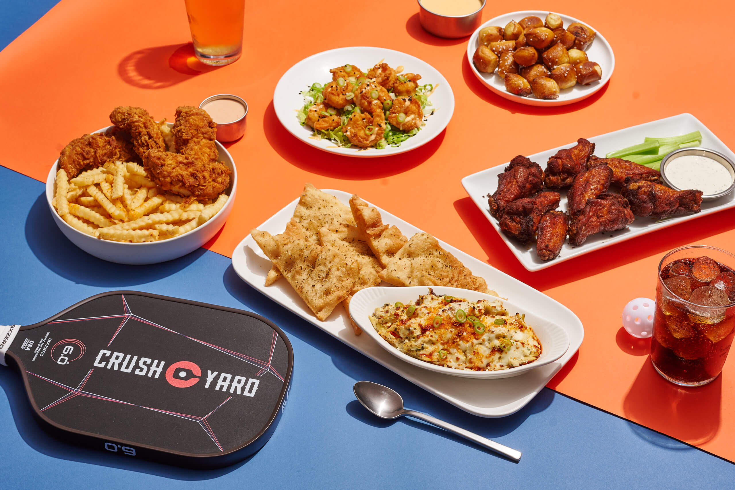 Assorted fried foods, chicken wings, flatbread, seasoned potatoes, and drinks are arranged on a table next to a Crush Yard pickleball paddle and ball—perfect for enjoying with the latest Nashville Deals.