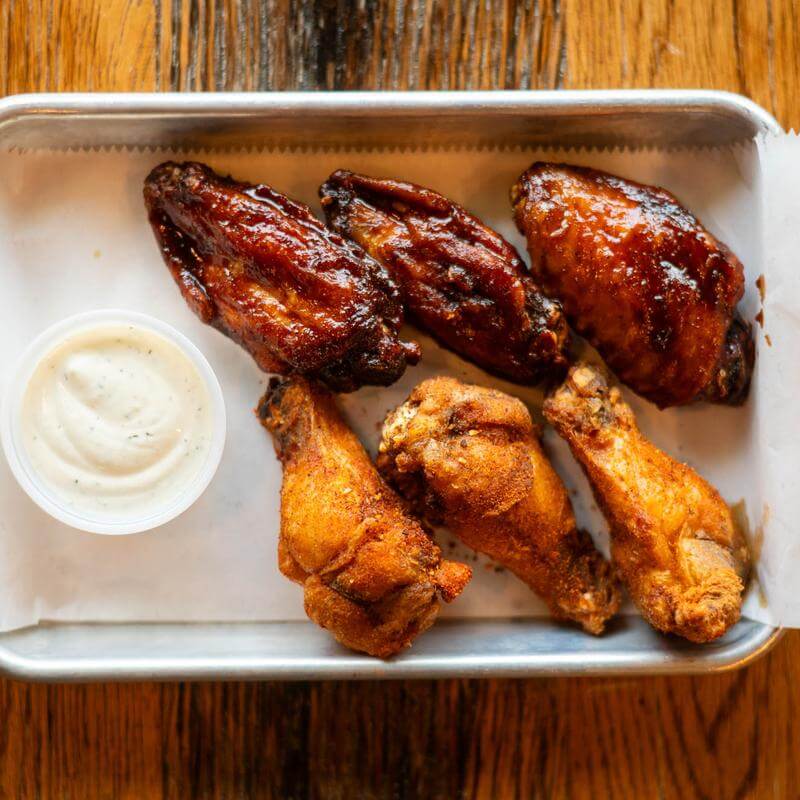 Nashville Steals, Deals & Announcements: June 2026 - 5 A metal tray with three sauced chicken wings, three fried drumsticks, and a cup of ranch dressing on parchment paper.