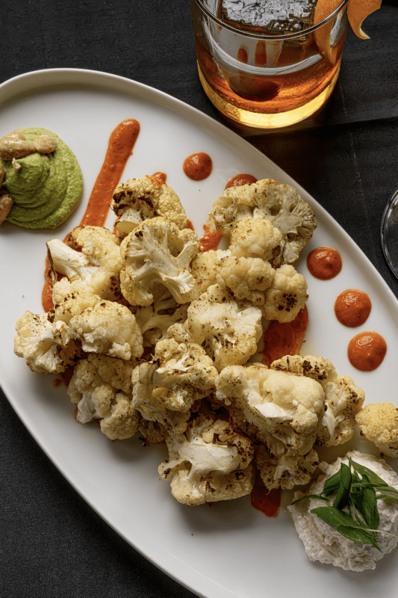 Nashville Steals, Deals & Announcements: June 2026 - 9 A white plate with roasted cauliflower, red sauce dots, green purée, a white dollop with herbs, and a drink with an orange peel garnish in the background.