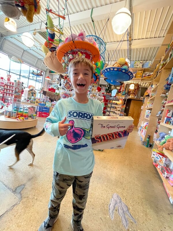 Your Guide to Nashville's Coolest Children's Stores - 3 Smiling child wearing a colorful hat and "Sorry!" shirt holds a "Sorry!" board game in a Nashville children's store.