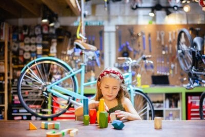 In a Nashville bicycle workshop, a young girl builds a structure with colorful wooden blocks at a table, surrounded by bikes and tools, creating an atmosphere reminiscent of playful children's stores.