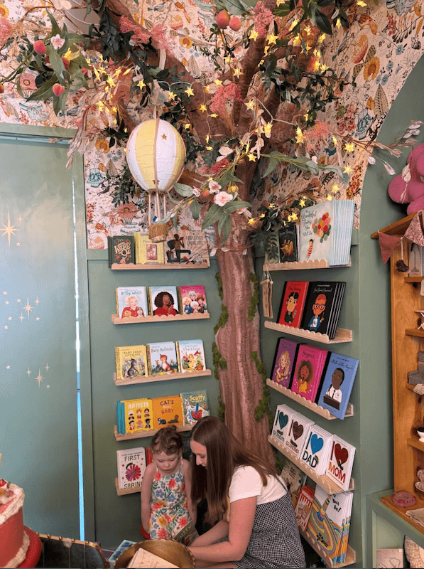 Your Guide to Nashville's Coolest Children's Stores - 9 A woman and a young girl sit reading in a colorful Nashville children’s bookstore corner, decorated with a large artificial tree, fairy lights, and shelves of picture books.