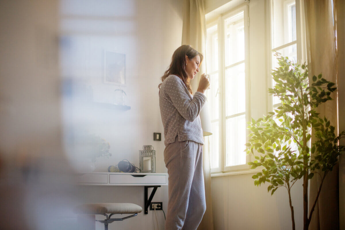 Tired, Wired, or Shut Down? Your Nervous System Wants Your Attention - 3 A woman in pajamas stands by a window, drinking from a mug in a sunlit room with a small plant and desk nearby, enjoying the quiet moment that helps her feel calmer and supports her nervous system.