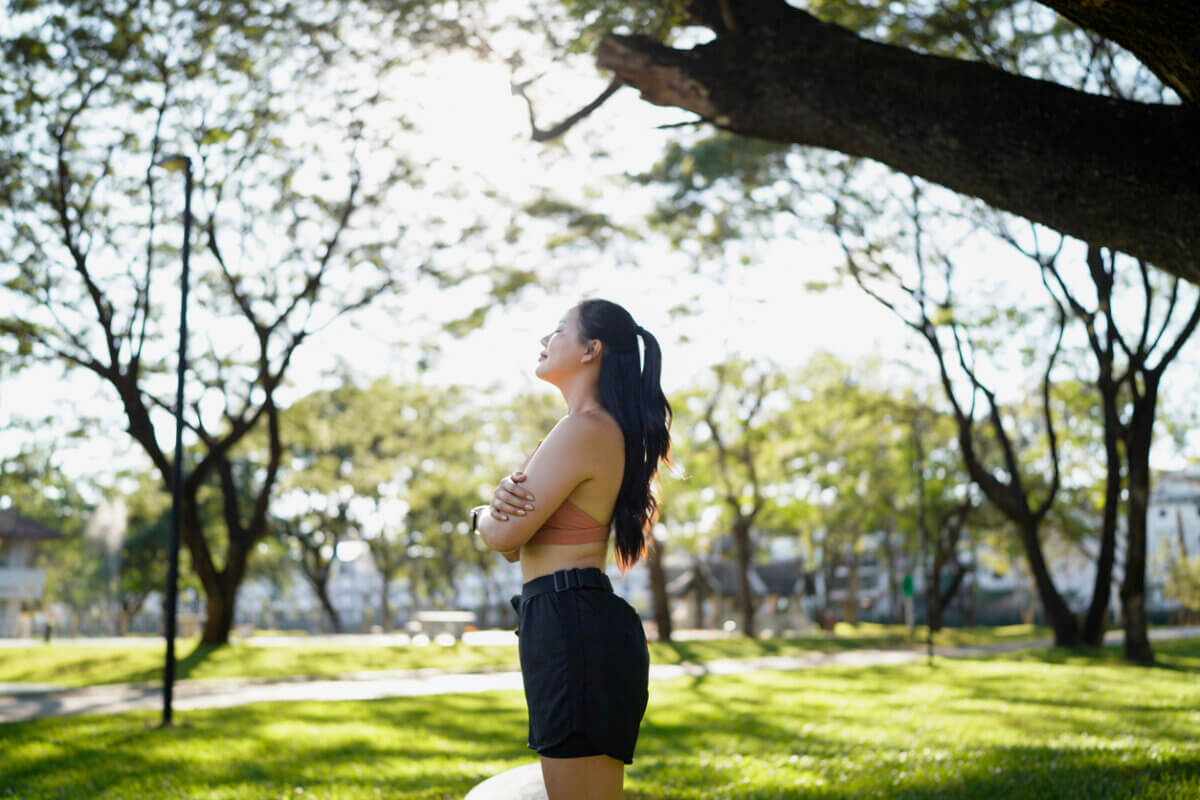 Tired, Wired, or Shut Down? Your Nervous System Wants Your Attention - 2 Woman standing outdoors in a park with arms crossed, looking up toward the sunlight, surrounded by green grass and treesβembracing nature to feel calmer and support her nervous system.