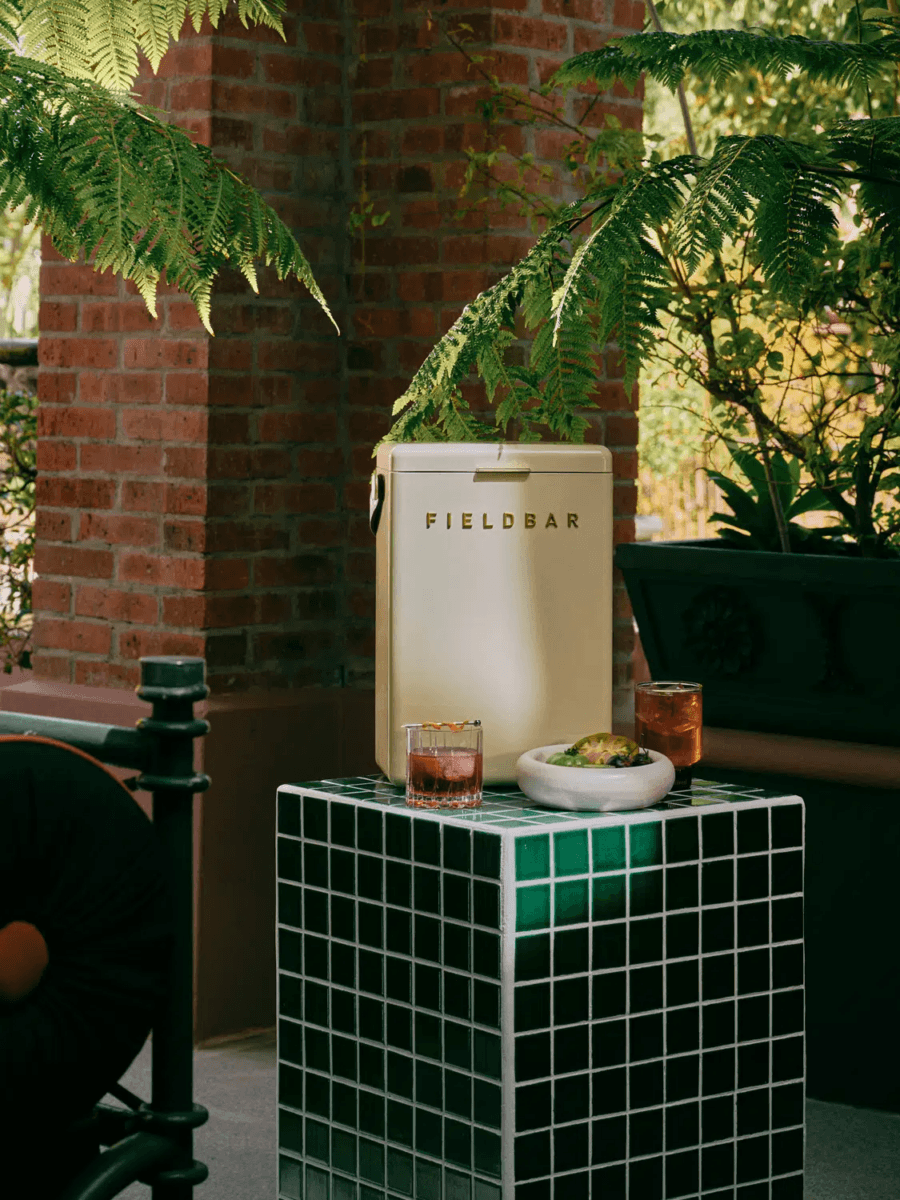 A Fieldbar cooler, an outdoor must-have, stands behind a green-tiled table holding two cocktails and a bowl of food, surrounded by greenery and brick columns.