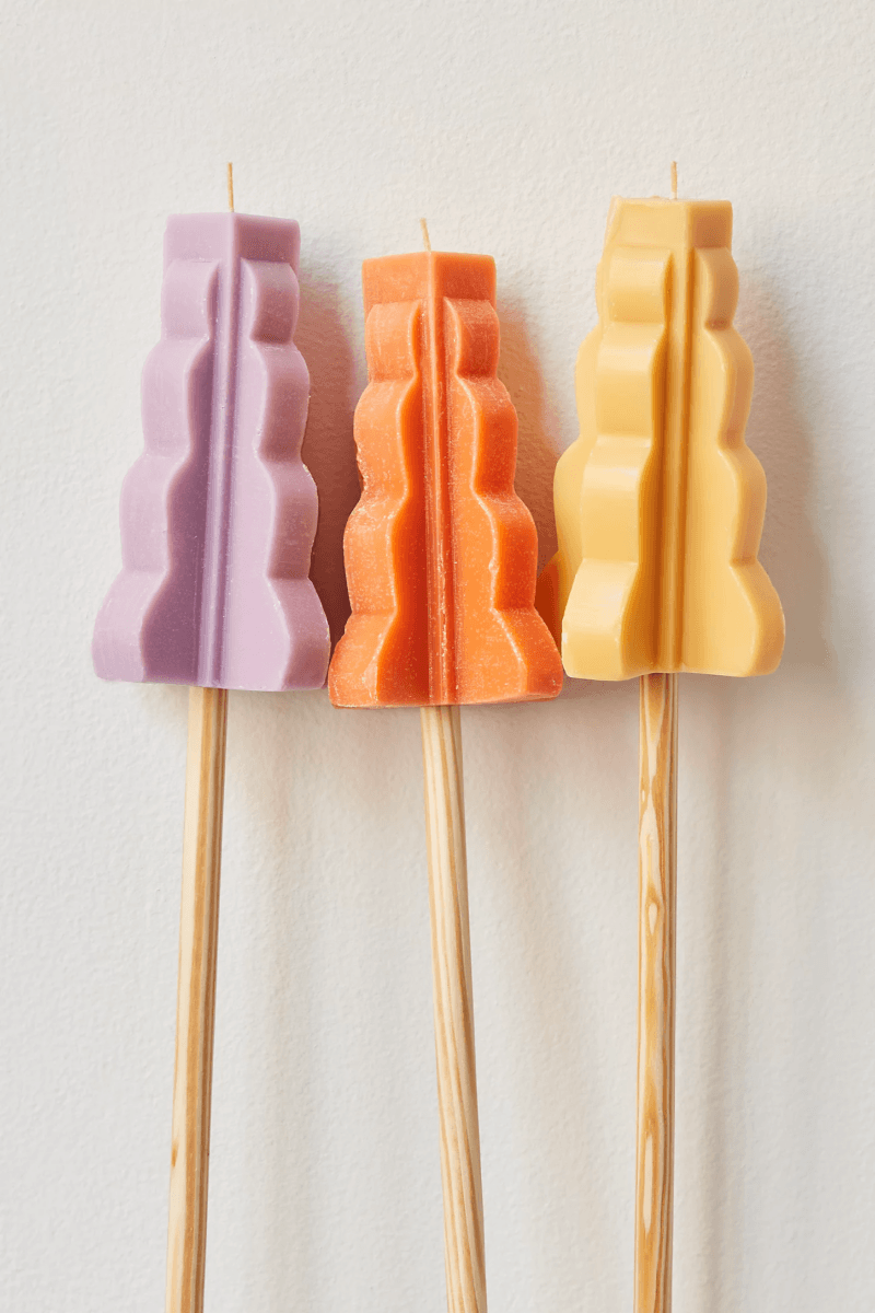 Three sculpted candles in purple, orange, and yellow, each attached to a wooden stick, make perfect outdoor entertaining essentials, displayed against a plain white background.