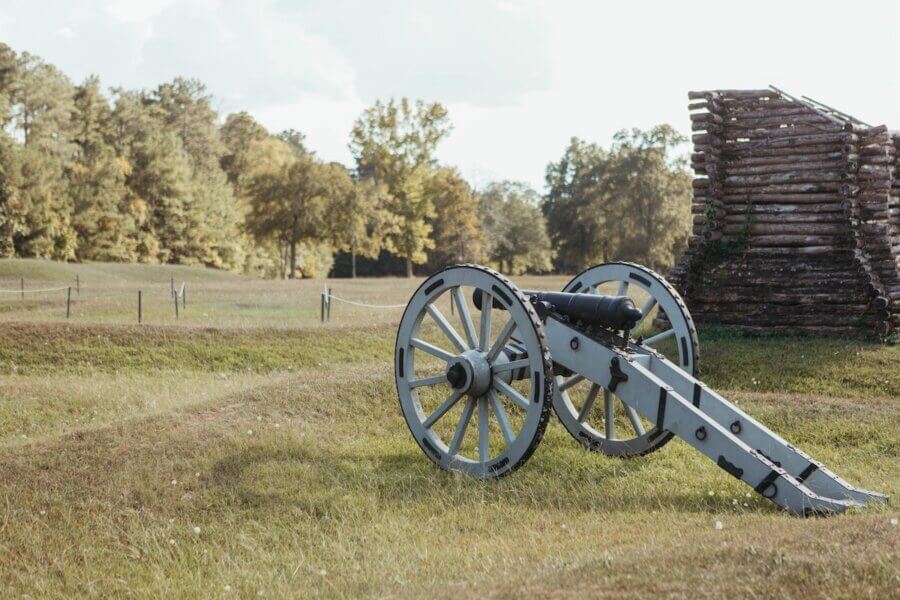 The Top Outdoor Adventures in South Carolina - 3 Historic cannon on a grassy field near a wooden log structure, with trees and a fence in the background under a partly cloudy skyβan iconic scene for South Carolina outdoor adventures.