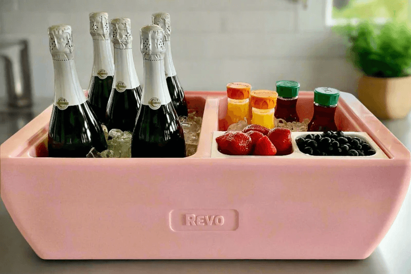 A pink Revo cooler filled with ice holds six bottles of sparkling wine, assorted juice bottles, strawberries, and blueberries—outdoor entertaining made easy on your kitchen counter.