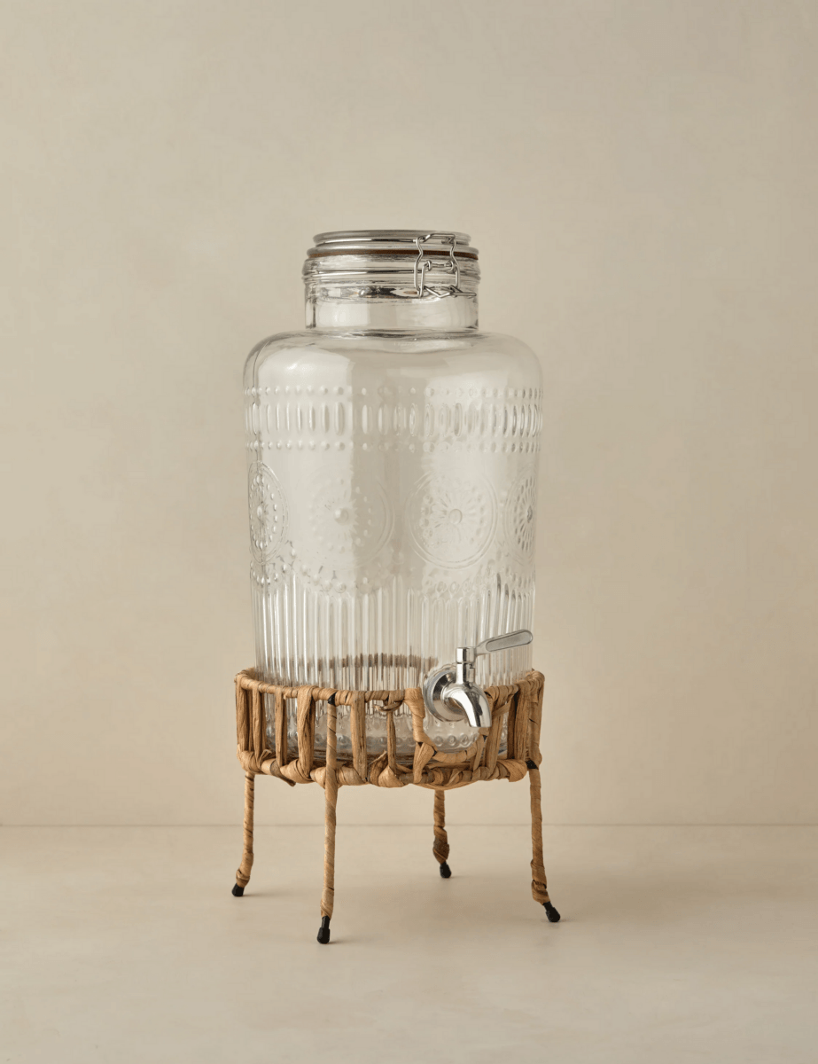 A clear glass beverage dispenser with a metal spigot and patterned design sits on a woven rattan stand—a must-have for stylish outdoor entertaining against a plain, light background.