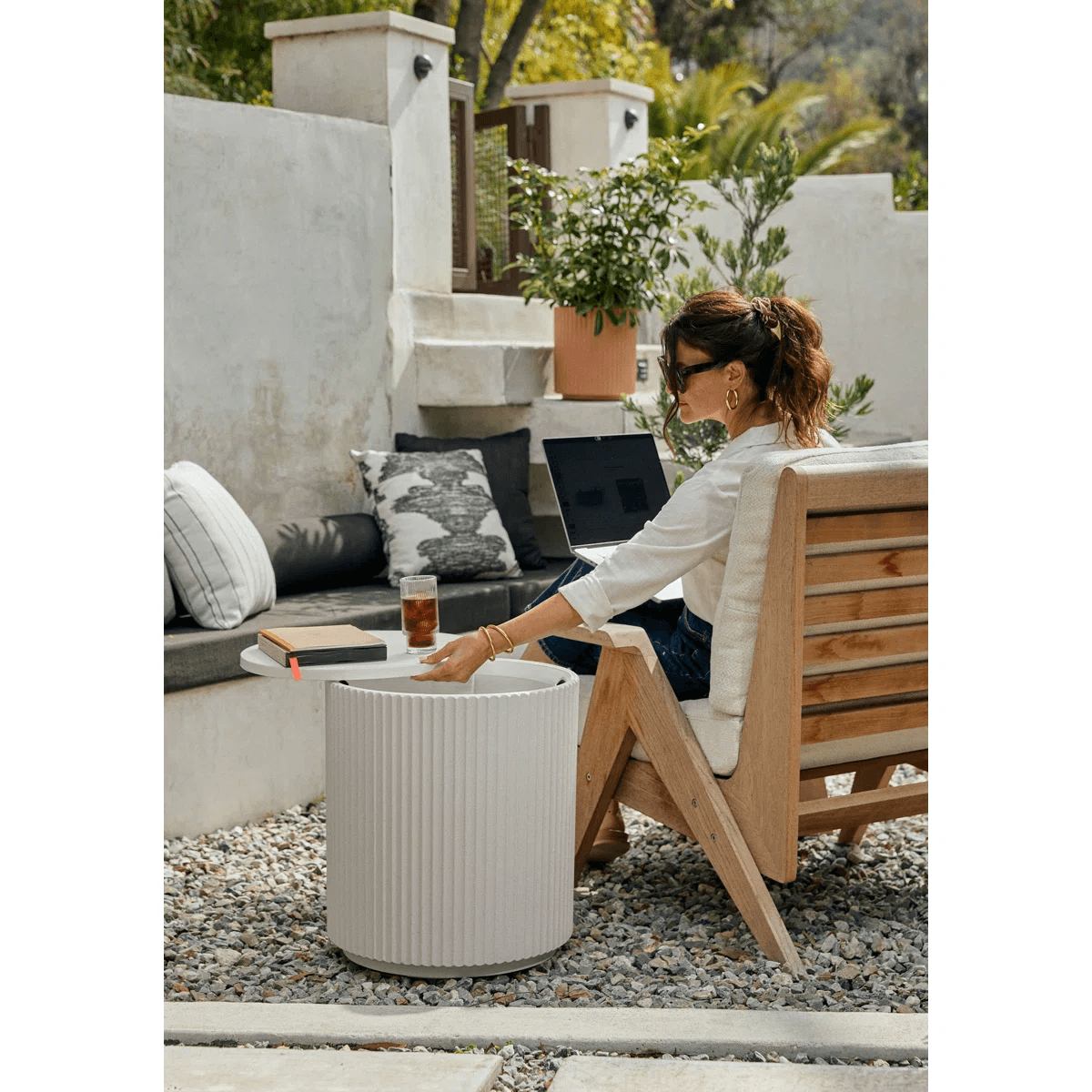 A woman sits on a wooden chair in an outdoor patio, using a laptop on her lap with a drink and book on a round white side table nearby.