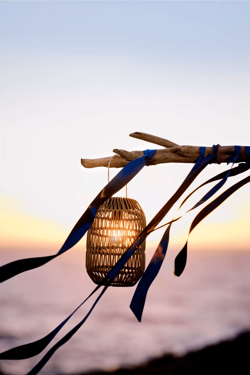 A woven lantern hangs from a branch decorated with blue ribbons, creating a must-have scene for outdoor entertaining, with the ocean and sunset in the background.