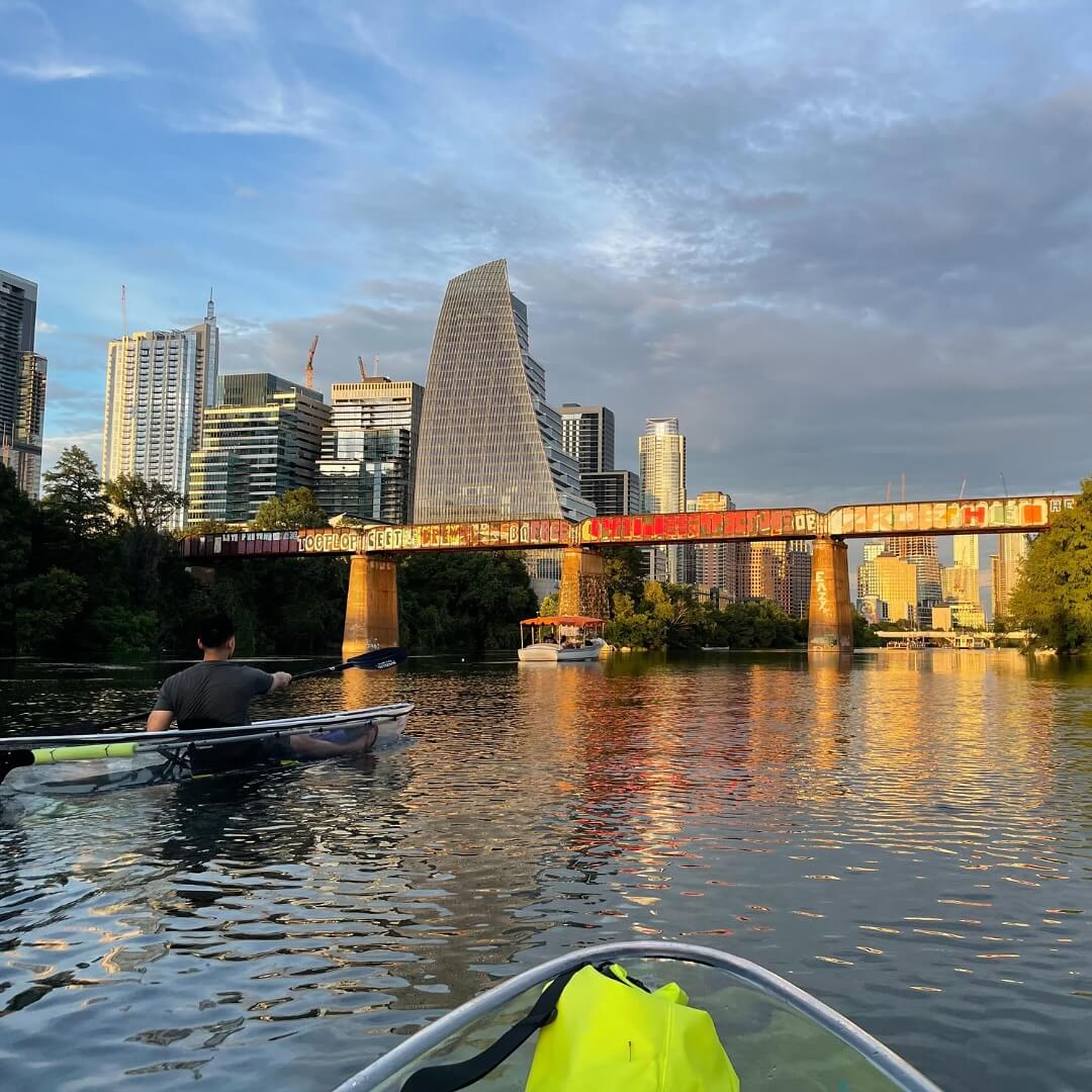 Epic Kayak Adventures in the South - 12 Two kayaks on a river glide beneath a graffiti-covered bridge, with the Austin skyline in the background at sunset.