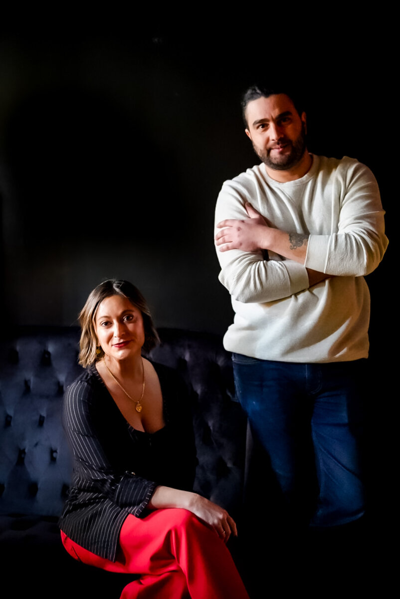 From Giving Shots to Serving Them: Meet Chattanooga's Cocktail Queen - 4 A woman sits on a dark couch while a man stands beside her with arms crossed, both facing the camera against a dark background, evoking the moody atmosphere of Calliope Chattanooga.