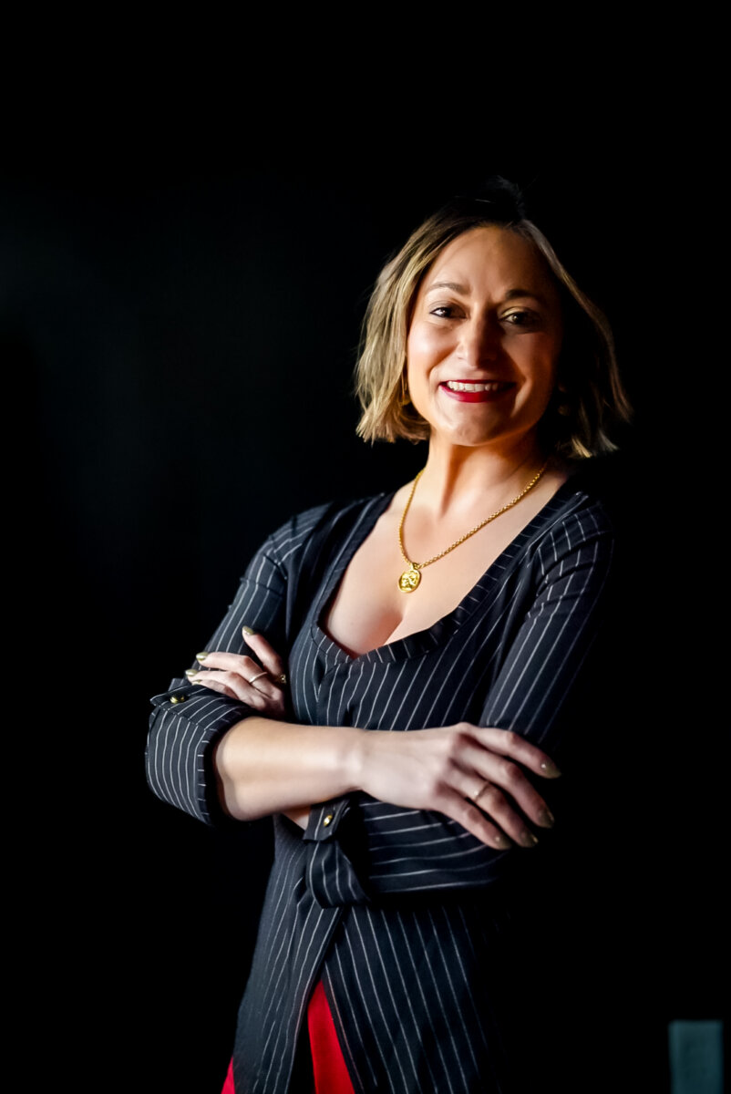 From Giving Shots to Serving Them: Meet Chattanooga's Cocktail Queen - 2 A woman entrepreneur with short hair wearing a pinstripe blazer, red pants, and a gold necklace stands with arms crossed, smiling confidently against a black background.