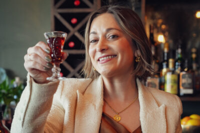 A woman behind a light-colored blazer holds up a small glass of dark red beverage, smiling, with bottles and fruit in the background, as if showcasing her choice from the beverage menu.