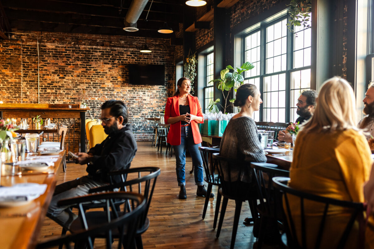From Giving Shots to Serving Them: Meet Chattanooga's Cocktail Queen - 3 A woman in a red blazer, known as the woman behind Calliope, stands near a group of people seated at a long wooden table in a restaurant with brick walls and large windows.