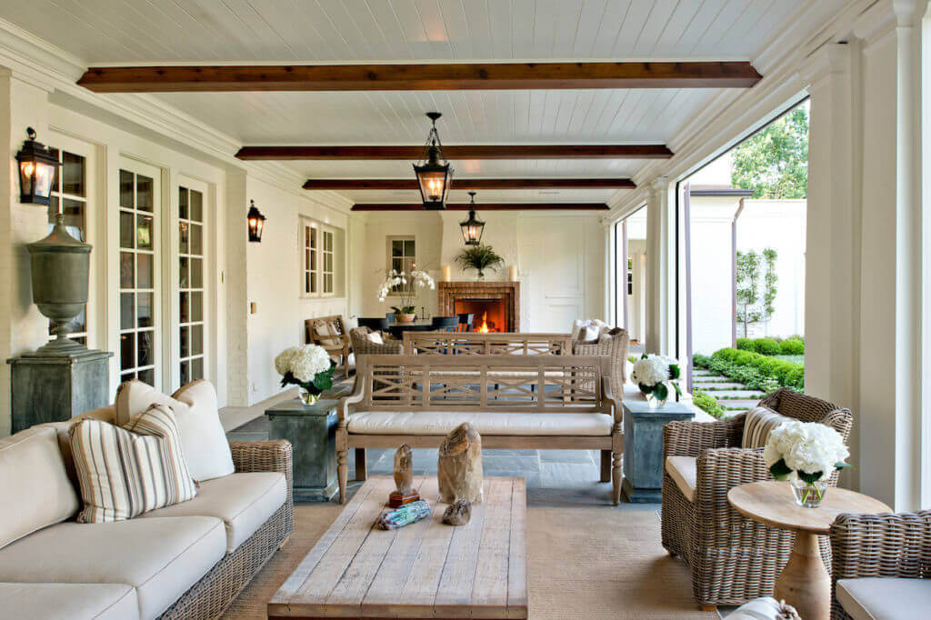 10 Designer Tips for Upgrading Your Outdoor Spaces - 11 A covered outdoor patio with wicker furniture, striped cushions, a wooden coffee table, potted flowers, and a fireplace at the far end offers expert tips for designing inviting outdoor spaces that overlook a landscaped garden.