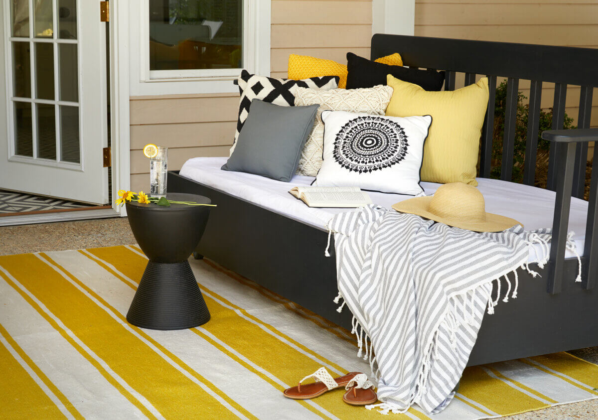 10 Designer Tips for Upgrading Your Outdoor Spaces - 7 A porch daybed with patterned pillows, a striped throw, an open book, a straw hat, a small table with a drink and flowers, and a yellow striped rug offers inspiring designing tips for creating beautiful outdoor spaces.