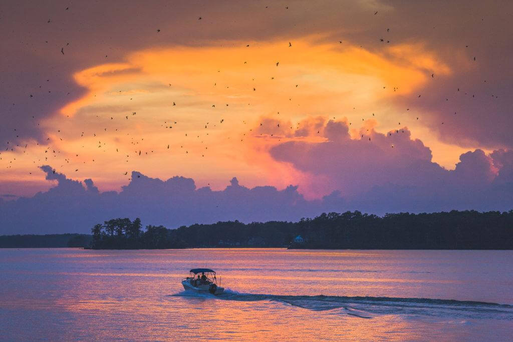The Best Summer Weekend Getaways in the South - 12 A motorboat glides across a calm lake at sunset in the South, perfect for summer weekend trips, with a dramatic orange and purple sky, silhouetted trees, and birds flying overhead.