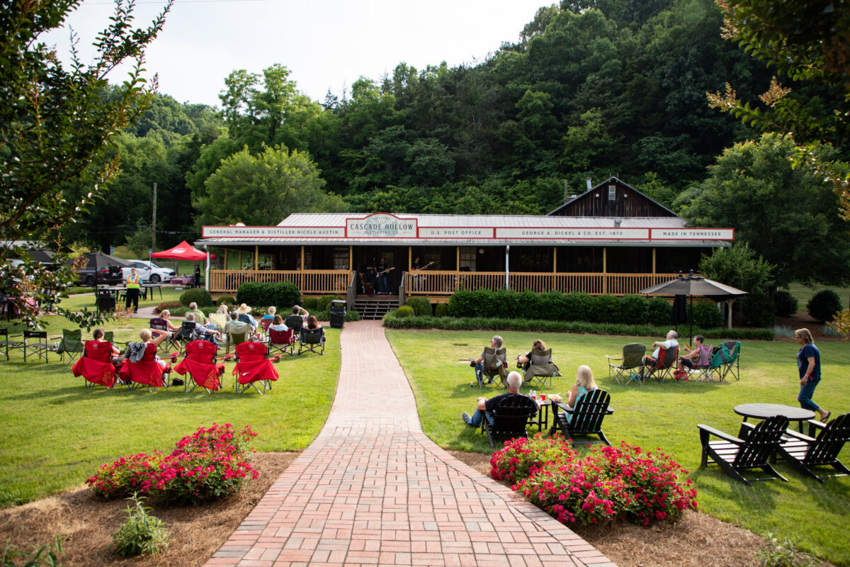 The Best Summer Weekend Getaways in the South - 14 People sit on lawn chairs and benches facing a building with a porch in a grassy outdoor setting, surrounded by trees and blooming flowers—an inviting scene perfect for weekend getaways in the South.