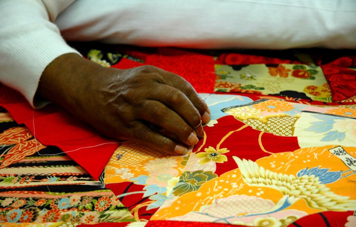The Best Summer Weekend Getaways in the South - 4 A person’s hand is sewing a colorful quilt featuring various patterned fabrics, including floral and crane designs, using a needle and white thread—perfect for cozying up after summer weekend trips in the South.