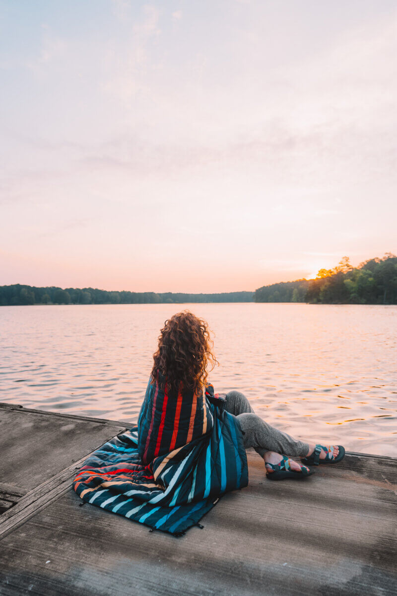 The Best Summer Weekend Getaways in the South - 11 Person with curly hair sits on a dock wrapped in a striped blanket, gazing over a calm lake at sunset—one of the best summer weekend trips in the South, surrounded by trees lining the tranquil shore.