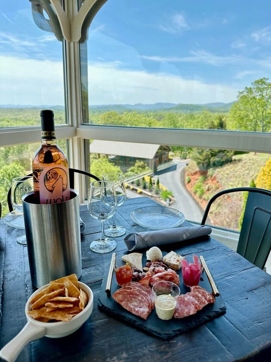 5 Fabulous Wine Weekends Around the South - 5 A charcuterie board, wine bottle in a cooler, and glasses are set on a table by a window overlooking a scenic landscape—perfect for relaxing during Southern wine trips or unforgettable wine weekends.