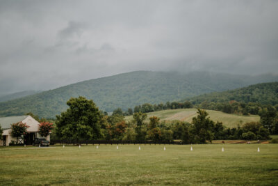 A large grassy field with scattered white cones, a barn-like building on the left, and tree-covered hills in the background under a cloudy South sky—perfect for wine weekends or leisurely wine tours.
