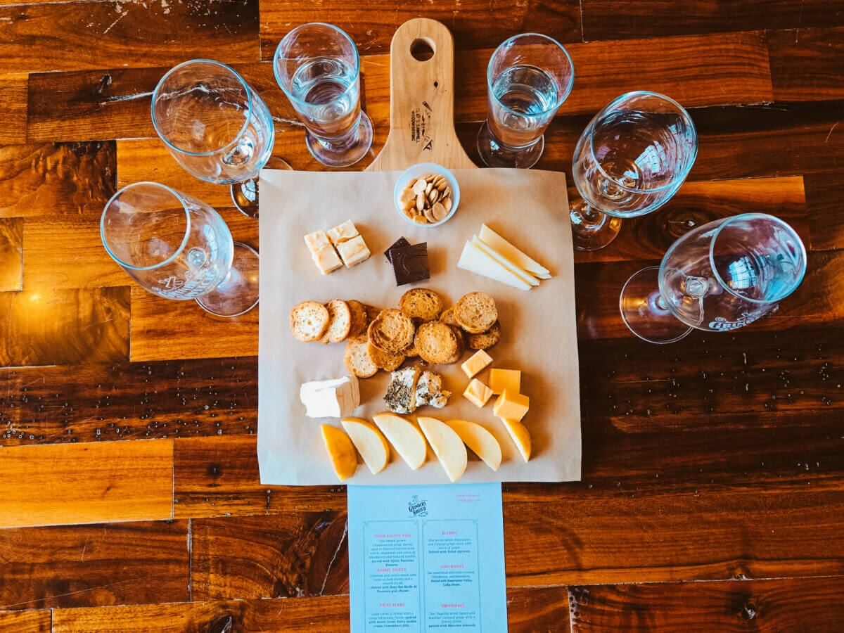 Need a Change of Scenery? Try These Summer Weekend Trips From Nashville - 4 A wooden board with assorted cheeses, crackers, nuts, and chocolate is surrounded by six empty wine glasses on a wooden table with a menu beneath—perfect for Nashville summer weekend trips or relaxing weekend getaways.