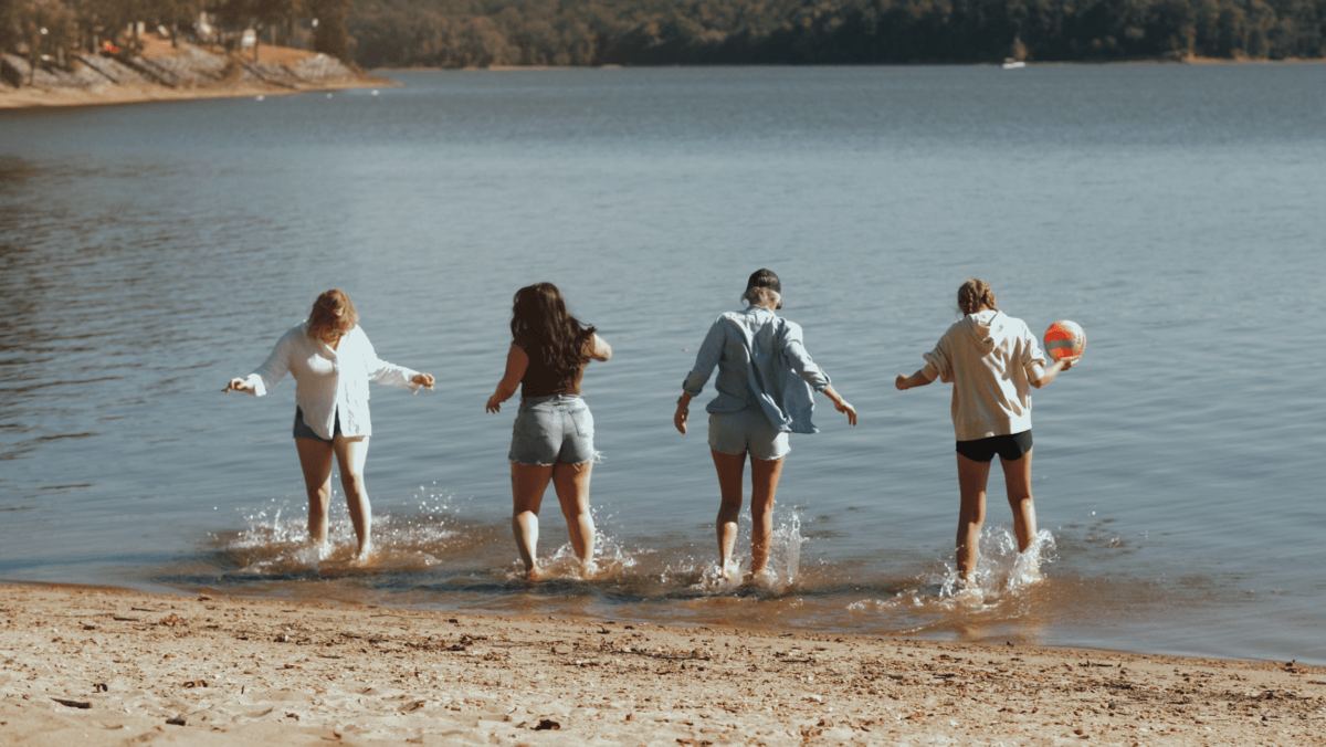 Need a Change of Scenery? Try These Summer Weekend Trips From Nashville - 10 Four people stand at the edge of a lake, splashing water with their feet during one of their weekend trips from Nashville. One person holds a colorful beach ball, with trees and a serene shoreline in the background.