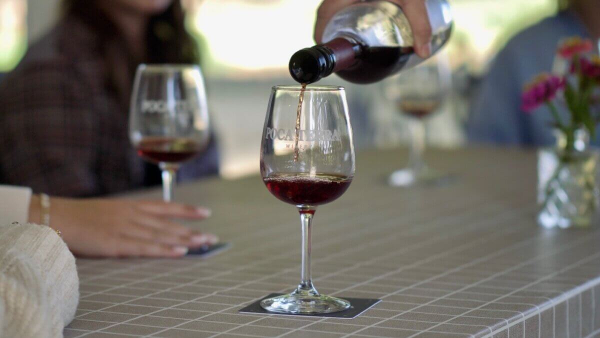 Need a Change of Scenery? Try These Summer Weekend Trips From Nashville - 11 A person pours red wine into a wine glass on a checkered tablecloth, capturing the relaxed vibe often found during weekend trips from Nashville, with other wine glasses and people enjoying themselves in the background.