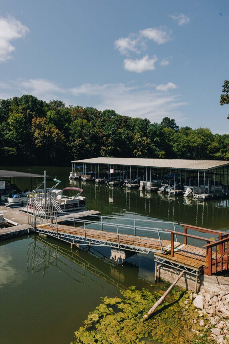 Need a Change of Scenery? Try These Summer Weekend Trips From Nashville - 2 A dock with boats on a calm lake, surrounded by trees under a partly cloudy sky. Algae is visible in the water near the dock, making it a peaceful spot for relaxing day trips from Nashville.