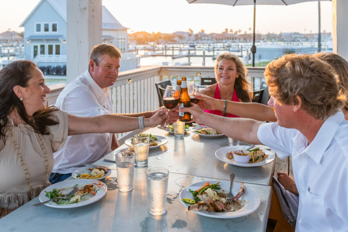 How to Eat, Shop & Play Your Way Through Texas - 6 Five people seated at an outdoor table by the water clink their drinks together over plates of food at sunset—an unforgettable way to eat in Texas.