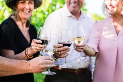 Four adults smiling and clinking glasses of red and white wine outdoors, surrounded by greenery, celebrate the good life—just another reason to eat in Texas and savor unforgettable moments together.