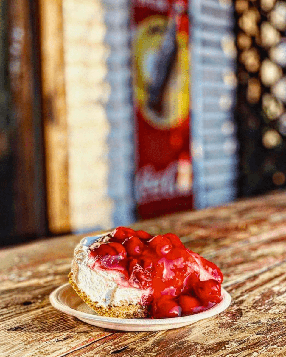 How to Eat, Shop & Play Your Way Through Texas - 11 A slice of cheesecake topped with cherry sauce on a disposable plate, placed on a rustic wooden table.