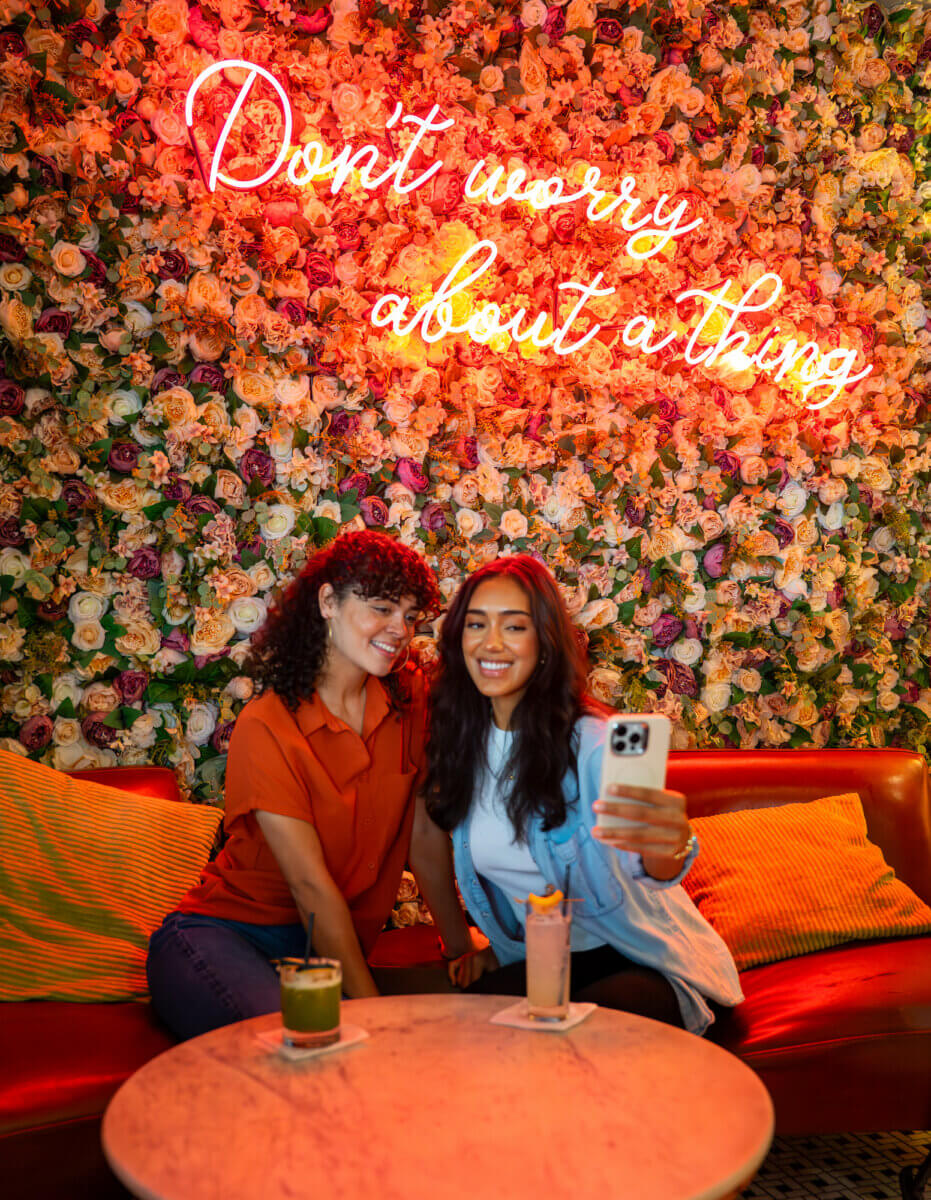 How to Eat, Shop & Play Your Way Through Texas - 9 Two women sit on a red couch taking a selfie in front of a flower-covered wall with a neon sign that reads "Don't worry about a thing," enjoying drinks as they Eat in Texas.