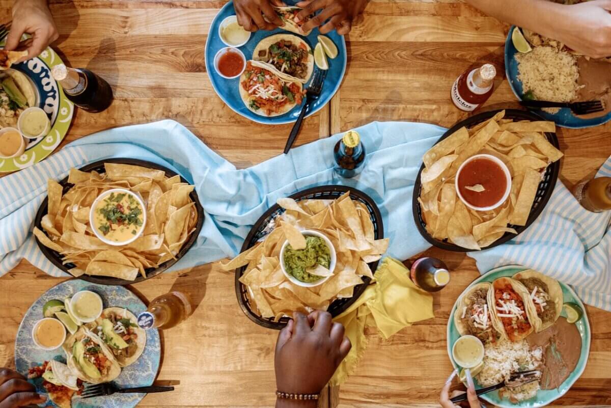 How to Eat, Shop & Play Your Way Through Texas - 2 Overhead view of people sharing plates of tacos, tortilla chips with salsa and guacamole, and drinks on a wooden table with a blue cloth.
