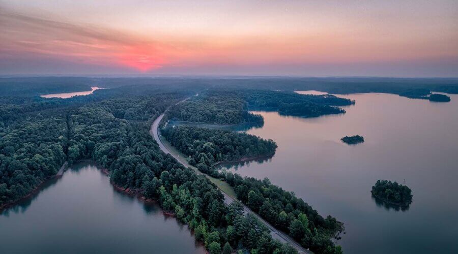 The Top Outdoor Adventures in South Carolina - 2 Aerial view of a winding road cutting through dense forest along the edge of a large lake at sunsetβperfect for South Carolina outdoor adventuresβwith calm water and a pink sky.
