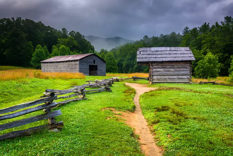 This Quiet Town is the Smokies' Best-Kept Secret - 7 A dirt path winds between two old wooden cabins in a quiet town, surrounded by split rail fences and green fields, with forested Smokies hills and a cloudy sky in the background.