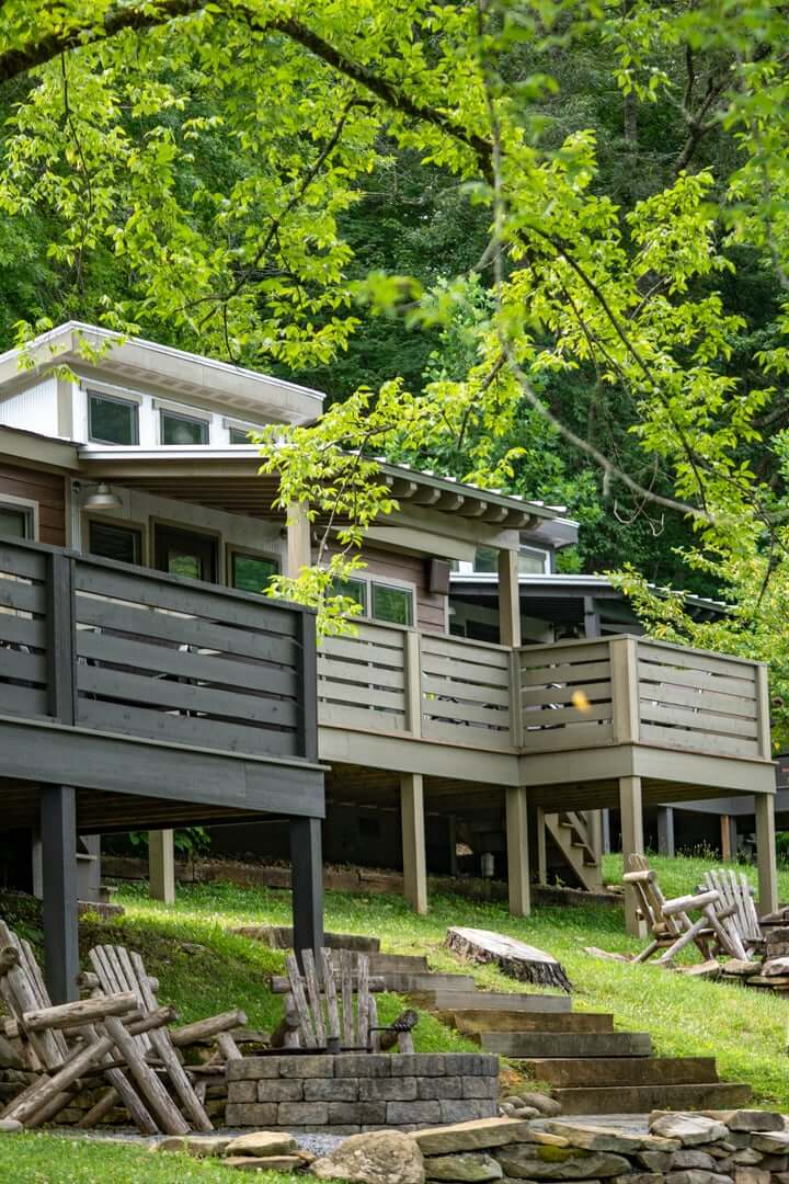 This Quiet Town is the Smokies' Best-Kept Secret - 2 A modern cabin with a large elevated deck sits among green trees in a quiet town near the Smokies, with wooden chairs and stone steps on the grassy lawn below.