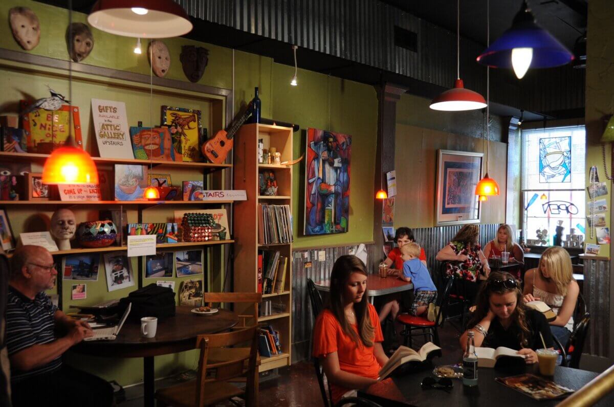 Why Vicksburg Might Be Mississippi's Most Unexpected Art Destination - 6 People reading and working at tables in a cozy, eclectic café in Vicksburg, Mississippi—an art destination filled with books, artwork, masks, and hanging lights.