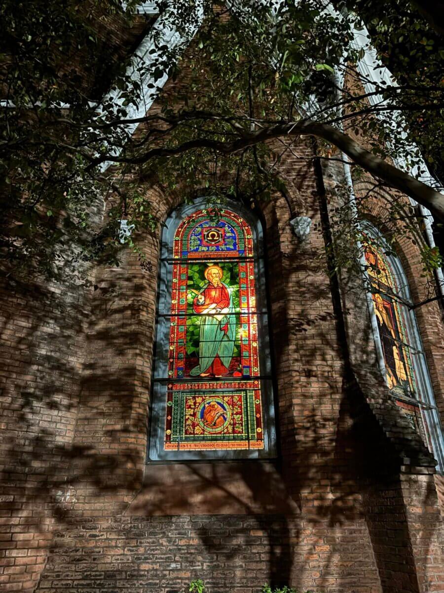 Why Vicksburg Might Be Mississippi's Most Unexpected Art Destination - 7 Stained glass window on a brick church wall in Vicksburg, Mississippi, illuminated at night, with tree branches casting shadows—an art destination for those who appreciate historic beauty.