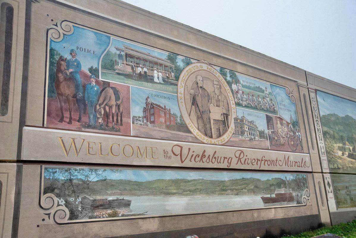 Why Vicksburg Might Be Mississippi's Most Unexpected Art Destination - 3 A mural on a concrete wall in Vicksburg displays historic scenes and figures, with text reading "Welcome to the Vicksburg Riverfront Murals," marking it as a standout Mississippi art destination.