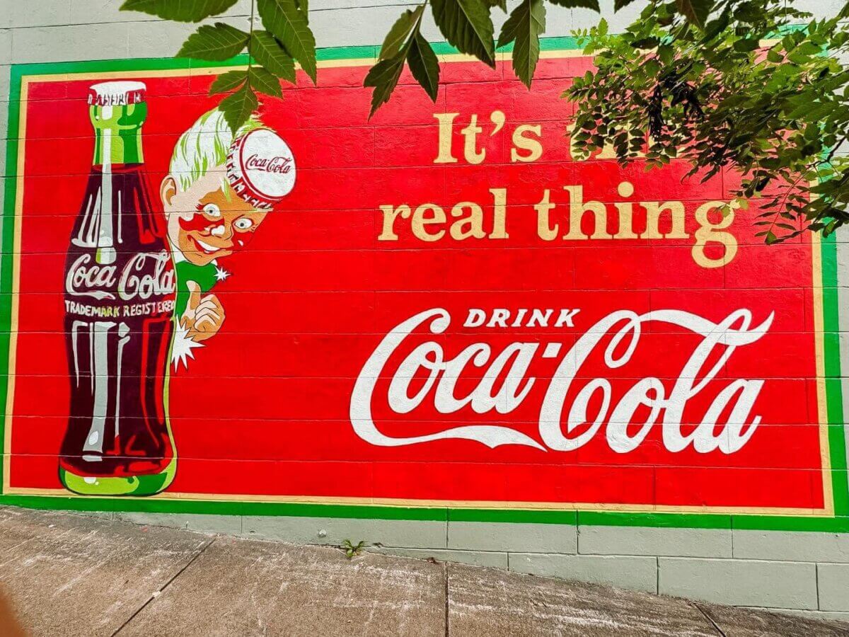 Why Vicksburg Might Be Mississippi's Most Unexpected Art Destination - 5 A mural on a wall in Vicksburg features a Coca-Cola bottle, the Coca-Cola logo, a cartoon character, and the slogan "It's the real thing. Drink Coca-Cola," making it a vibrant Mississippi art destination. Leaves are visible at the top.