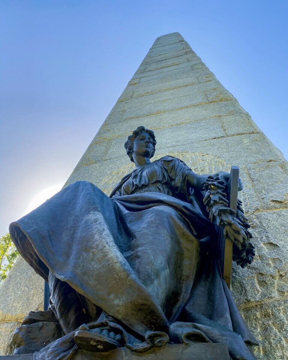 Why Vicksburg Might Be Mississippi's Most Unexpected Art Destination - 2 A stone obelisk rises behind a seated bronze statue of a woman holding a wreath, set beneath the clear blue sky of Vicksburg, making it an impressive art destination in Mississippi.