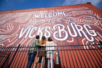 Three people stand with arms around each other, facing a large mural on a red brick wall that says "Welcome to Vicksburg," highlighting this Mississippi city as an art destination.